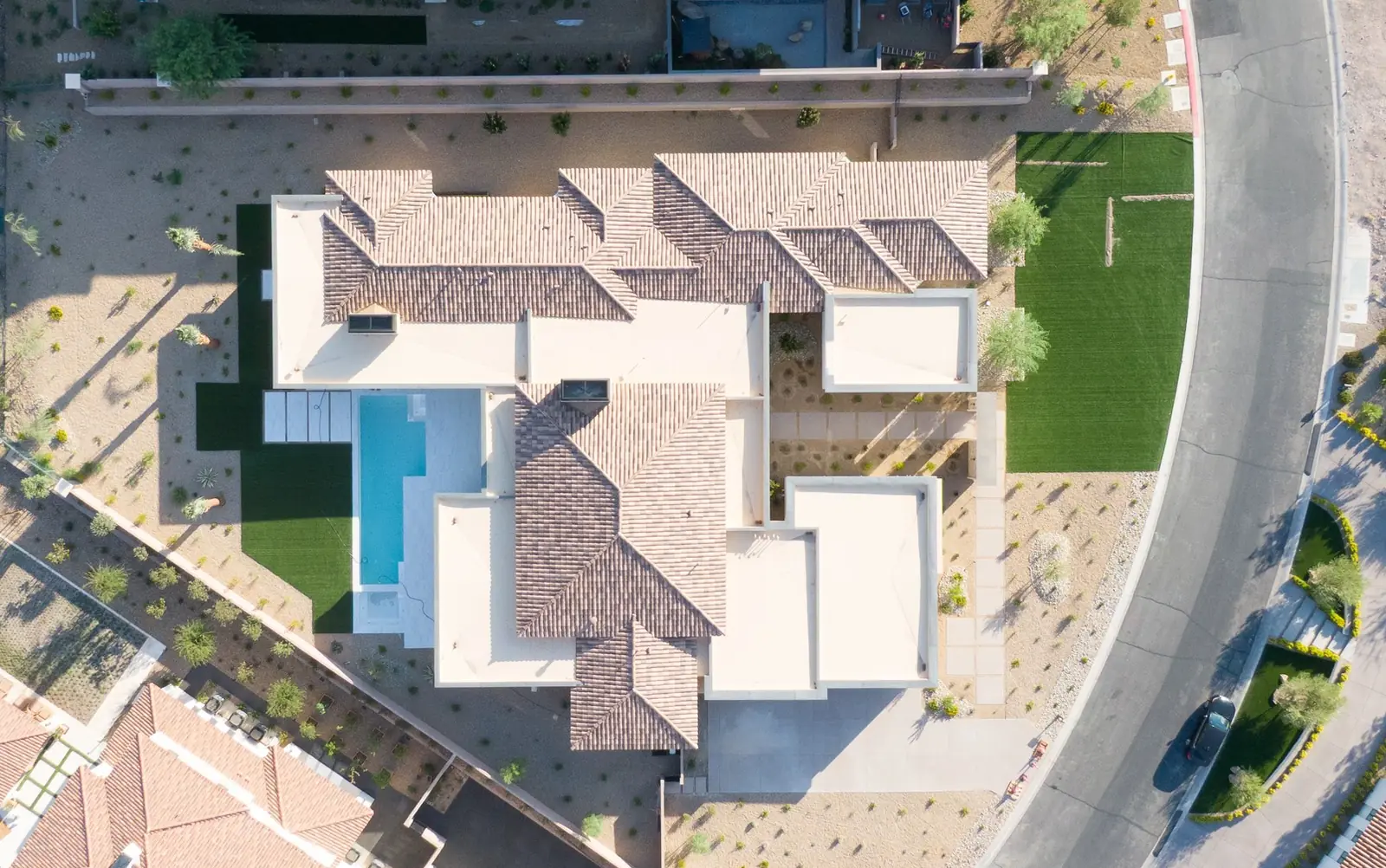 Straight down aerial of the house showing the tile roof, the covered patio, the pool with its wet deck, the turf strips and the street at the edge of the lot